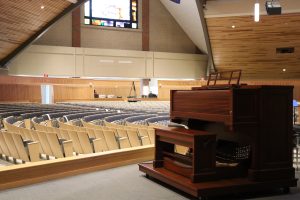 A spacious church auditorium with wooden pews aligned in rows, a large pipe organ in the foreground, and a video screen on the far wall displaying an image. The ceiling and walls are lined with wood paneling, and several religious inscriptions are carved into the walls.