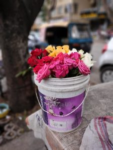 Bucket of vibrant roses in different colors, placed outdoors on a roadside surface, with blurred vehicles and a tree in the background.