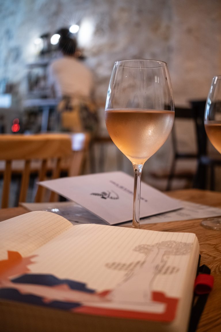 A glass of rosé wine sits on a wooden table next to an open notebook with sketches and writing. In the background, a blurred figure stands in a softly lit room with a warm, relaxed atmosphere.