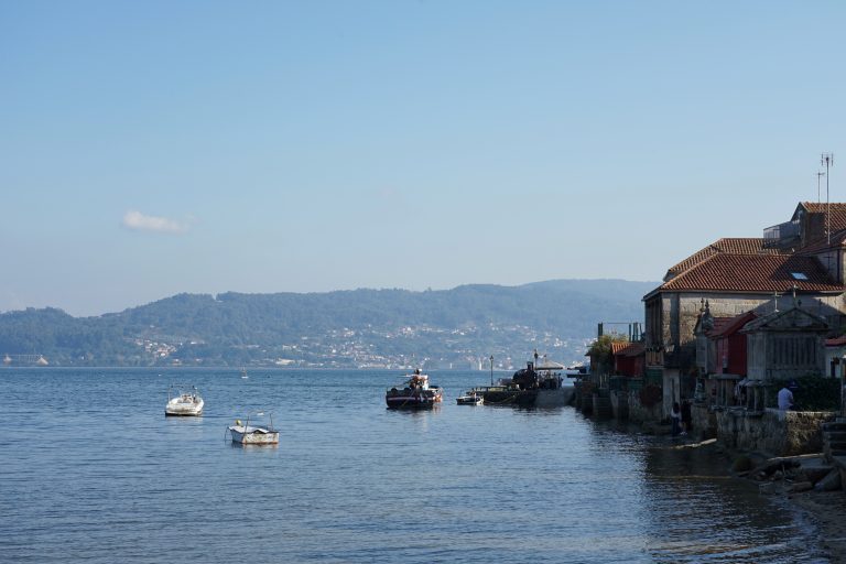 A scenic view of a coastal village with several small boats floating on calm waters. Traditional stone buildings with red-tiled roofs line the right side of the image, while a forested hillside is visible in the background across the bay.