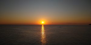 The image depicts a sunset in shades of orange over the sea. The surface of the water reflects the colors of the sky, creating playful glimmers on the waves.