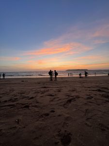 The image captures a serene beach at sunset, with a vibrant sky of soft pink and orange hues blending into blue. Silhouetted figures stand on the shoreline, adding a peaceful, contemplative feel.