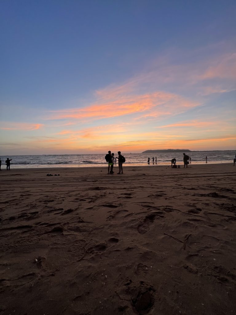 The image captures a serene beach at sunset, with a vibrant sky of soft pink and orange hues blending into blue. Silhouetted figures stand on the shoreline, adding a peaceful, contemplative feel.