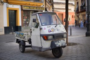 A small, white, three-wheeled delivery vehicle with graffiti parked on a cobblestone street in an urban area. The background features colorful buildings and a shop with a sign in Spanish.