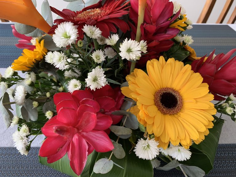 A vibrant floral arrangement featuring a yellow gerbera daisy, red and orange flowers, small white blossoms, and lush green leaves on a striped tablecloth background.