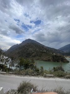 The photo shows a mountain with scattered small houses, rising above a greenish-blue lake or river. In the foreground, a road runs alongside bushes and small trees. The sky is partly cloudy, with patches of blue visible.