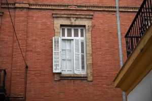 View larger photo: A red brick wall with a white window in the center, featuring open shutters. A balcony railing and part of a building edge are visible on the right side.