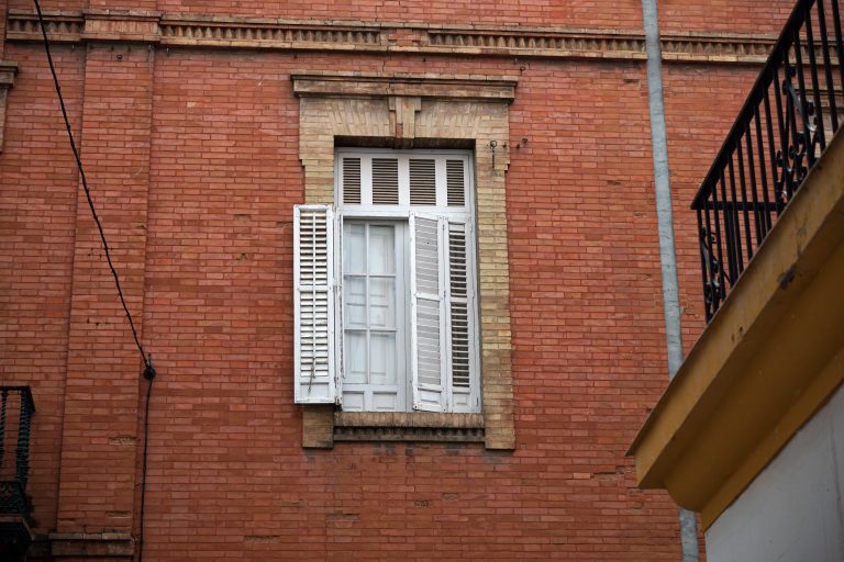 A red brick wall with a white window in the center, featuring open shutters. A balcony railing and part of a building edge are visible on the right side.