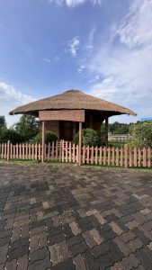 A small thatched-roof cottage labeled "Artists' Cottage," surrounded by greenery and a low wooden fence. The foreground features a paved stone walkway, and the sky is clear with a few clouds.