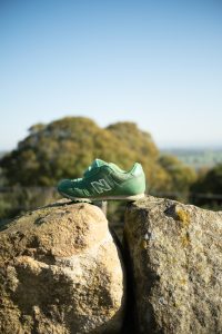 View larger photo: A small green New Balance shoe is perched on top of two large, weathered rocks. The scene is set against a backdrop of distant, blurred trees and a bright, clear blue sky, suggesting a peaceful, outdoor environment with a natural landscape.