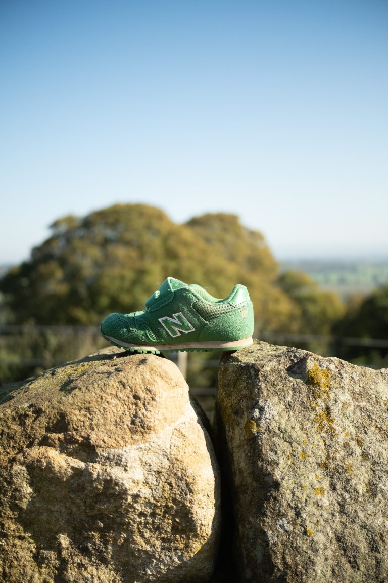 A small green New Balance shoe is perched on top of two large, weathered rocks. The scene is set against a backdrop of distant, blurred trees and a bright, clear blue sky, suggesting a peaceful, outdoor environment with a natural landscape.