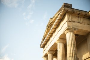 View larger photo: The upper section of a classical stone building with large, fluted columns and a detailed entablature, bathed in warm sunlight. The clear sky in the background enhances the grandeur and simplicity of the structure.