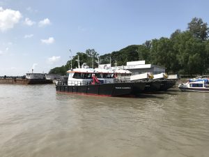 View larger photo: Boats in the Pasur River, Mongla, Myanmar. Trees along the banks and a blue sky overhead.