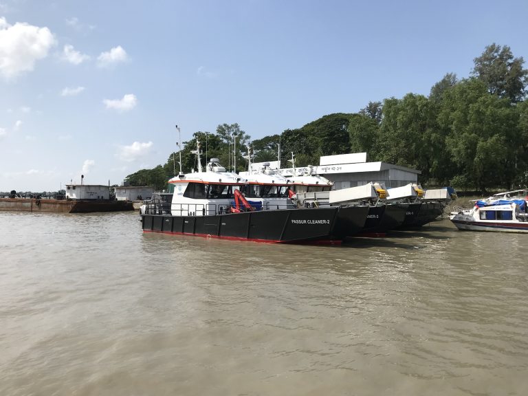 Boats in the Pasur River, Mongla, Myanmar. Trees along the banks and a blue sky overhead.