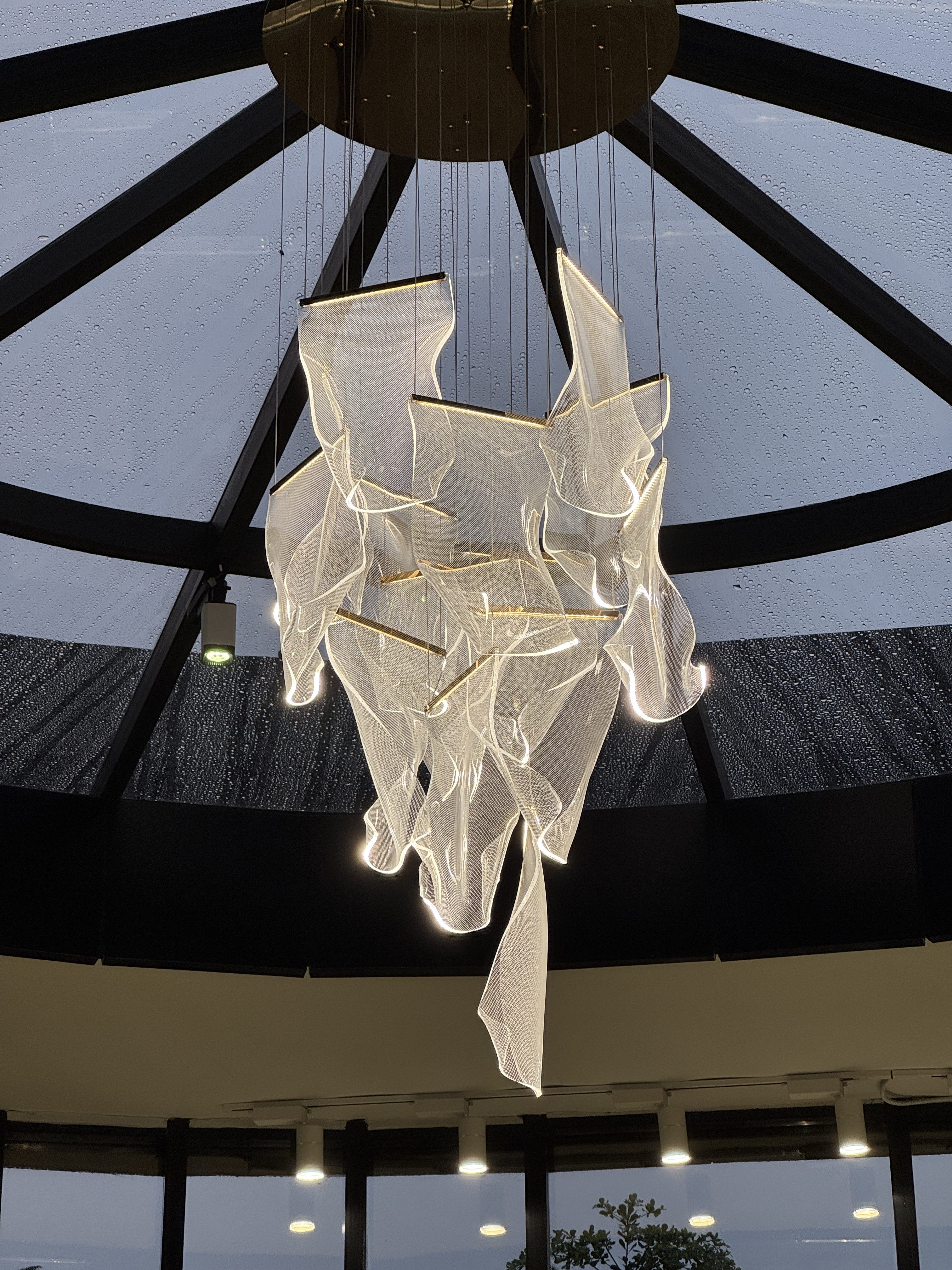 A modern, abstract chandelier made of translucent, flowing material with integrated lighting hangs from a ceiling beneath a geometric glass dome structure, visible during a rainy day with water droplets on the glass.