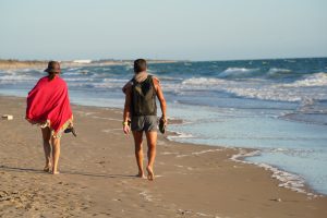 Two people walk barefoot along a sandy beach. One is wearing a hat and red cover-up, carrying sandals. The other has a backpack and sandals in hand, with a towel draped over their shoulder. 