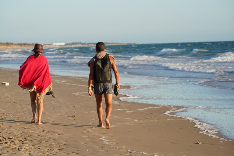 Two people walk barefoot along a sandy beach. One is wearing a hat and red cover-up, carrying sandals. The other has a backpack and sandals in hand, with a towel draped over their shoulder.