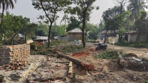 A village scene with scattered debris, fallen trees, and a broken house. A motorbike is parked on a dirt road, surrounded by lush greenery and small houses with tin roofs in the background.