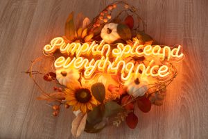 A decorative arrangement featuring a neon sign that reads 'Pumpkin spice and everything nice,' surrounded by artificial pumpkins, sunflowers, and autumn leaves. The glowing orange light casts a warm and cosy glow over the rustic wooden surface beneath it.