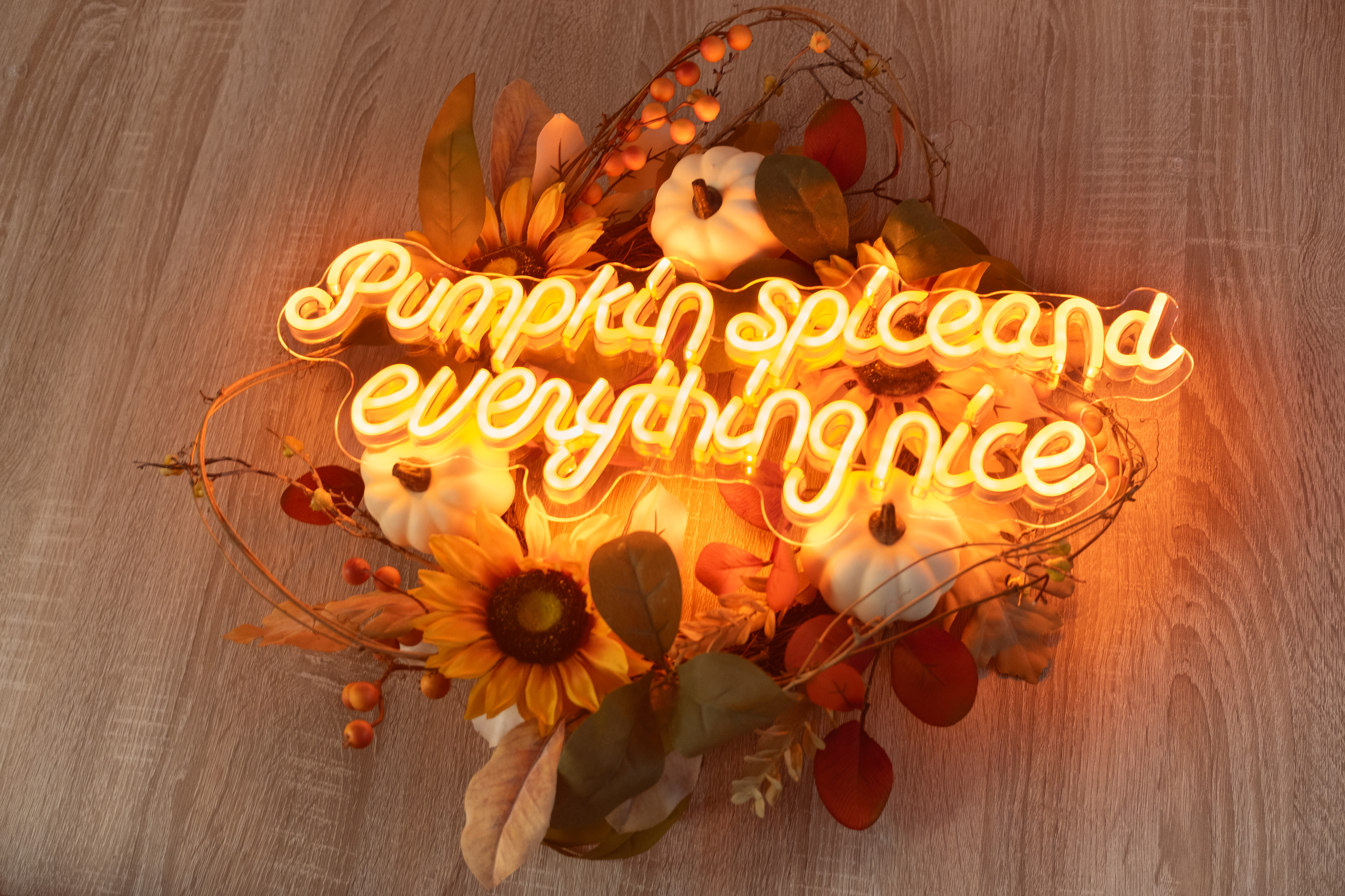 A decorative arrangement featuring a neon sign that reads 'Pumpkin spice and everything nice,' surrounded by artificial pumpkins, sunflowers, and autumn leaves. The glowing orange light casts a warm and cosy glow over the rustic wooden surface beneath it.