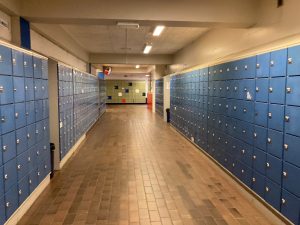 A school hallway with blue lockers on both sides of the hall. At the end of the hallways there are some green lockers.