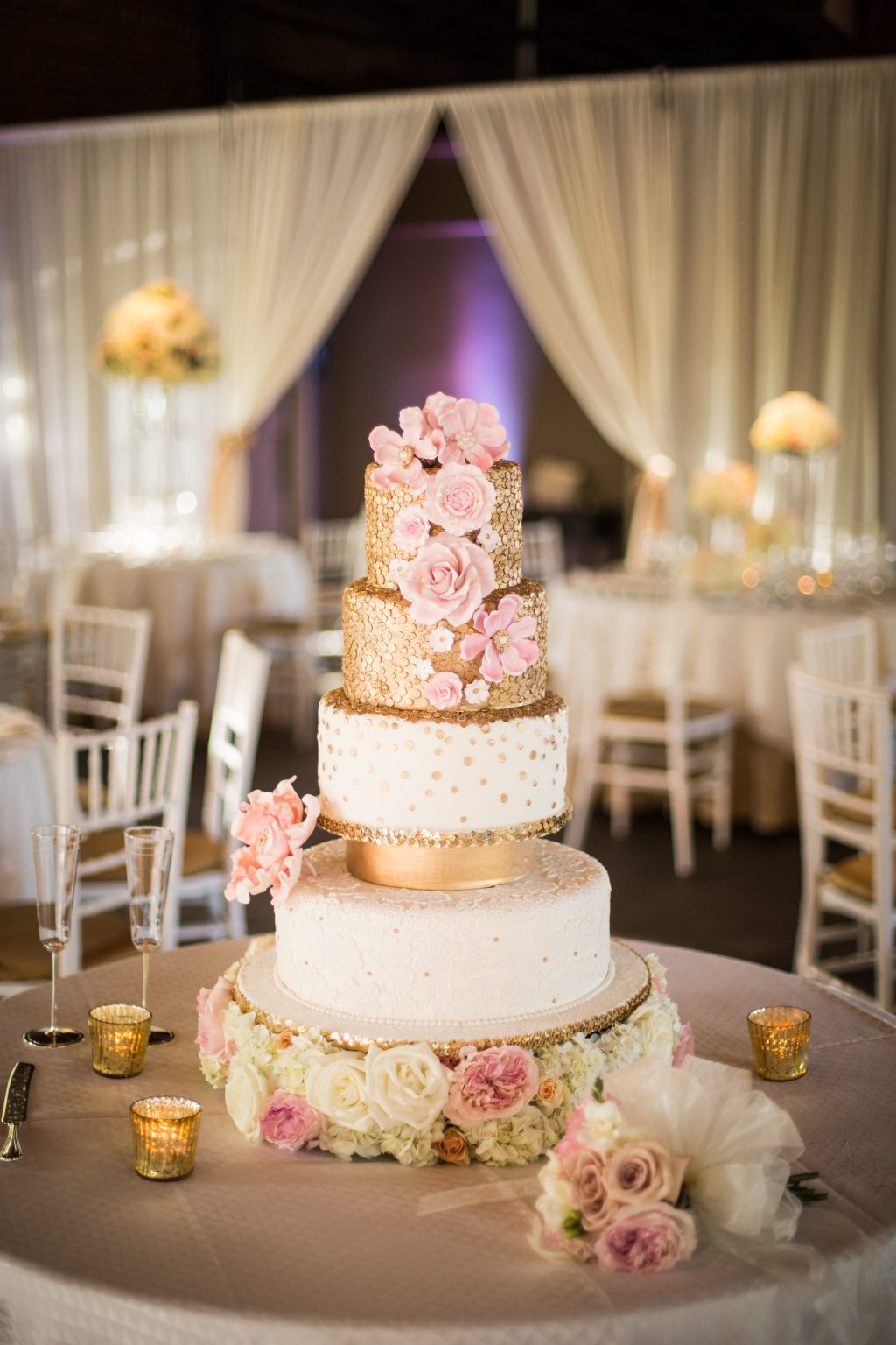 A four-tier wedding cake in white and gold with pink sugar roses, on a table ringed with fresh flowers and candles.