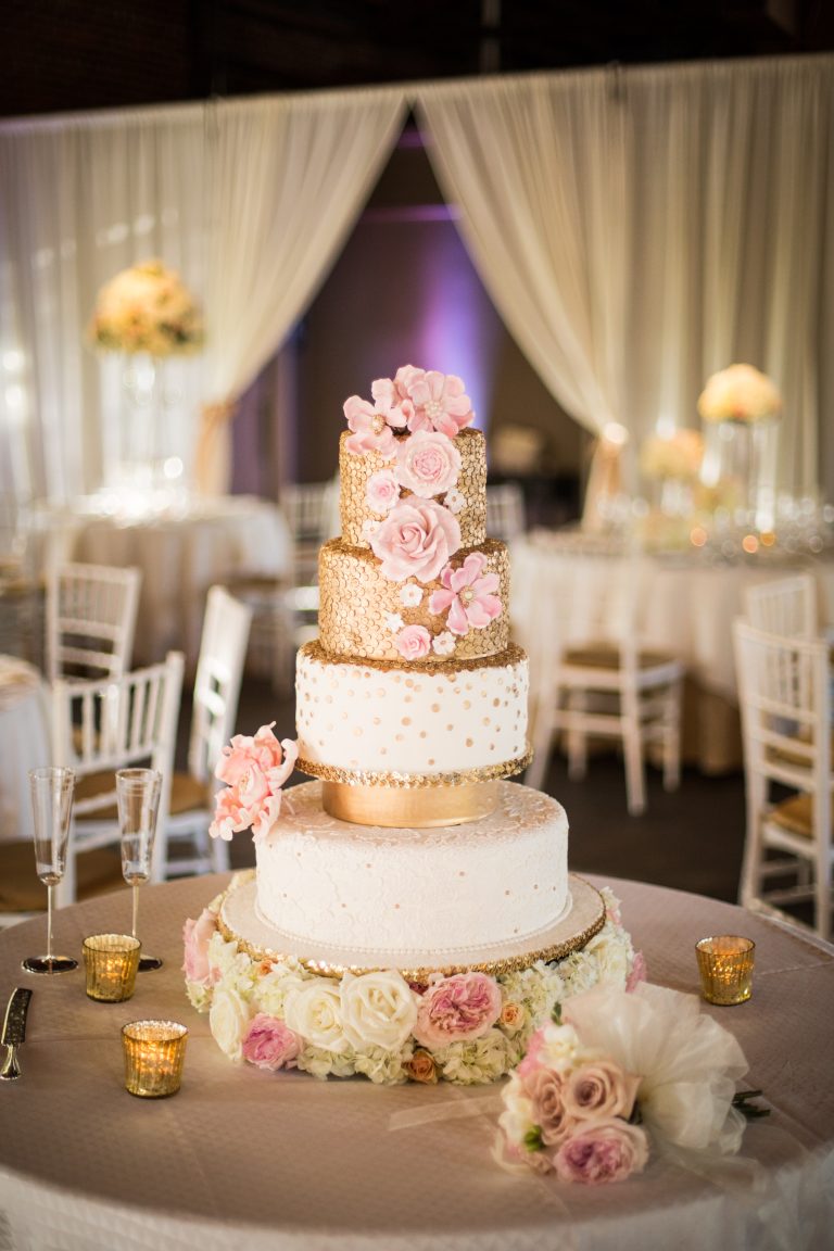 A gold and white tiered wedding cake with pink flowers.