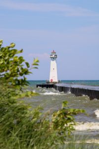 View larger photo: A lighthouse stands at the end of a pier on Lake Ontario as waves crash against the pier with a blue sky overhead and leaves framing it in the foreground.