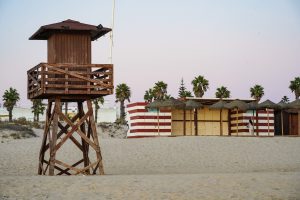 View larger photo: A wooden lifeguard tower stands on a sandy beach next to a structure with red and white stripes. In the background, there are palm trees and some buildings under a clear sky with a hint of pink at the horizon.