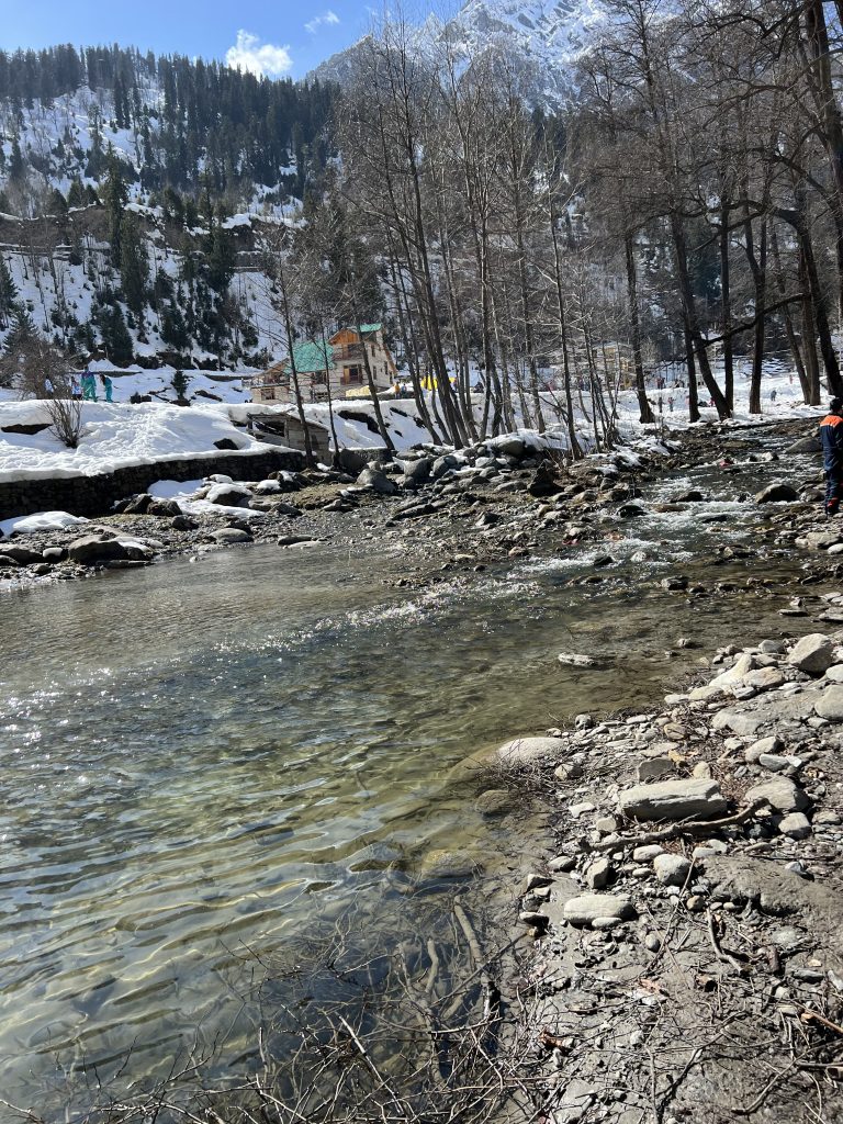 The image captures a serene winter landscape with a clear, shallow stream flowing over a rocky riverbank. Snow-covered ground surrounds the area, with bare trees in the foreground. In the background, snow-capped mountains and evergreen trees frame a few colourful buildings, likely part of a village.