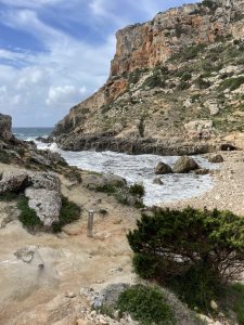 A rugged coastal landscape featuring a rocky shoreline and a steep cliff. The sea waves crash against the rocks, and the sky is partly cloudy. Vegetation, including shrubs and grass, grows among the rocky terrain. Cala Llucalari - Menorca