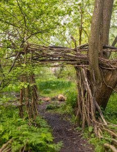 A rustic wooden archway made from intertwined branches and tree trunks stands along a narrow dirt path in a lush, green woodland. The dense foliage and natural materials blend seamlessly with the surrounding forest, creating a peaceful and secluded atmosphere.