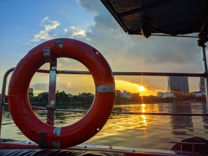 A bright orange lifesaver is affixed to the railing of a boat, with a scenic view of the sun setting over the horizon, casting a golden glow on the water and silhouetting distant city buildings against a partly cloudy sky.