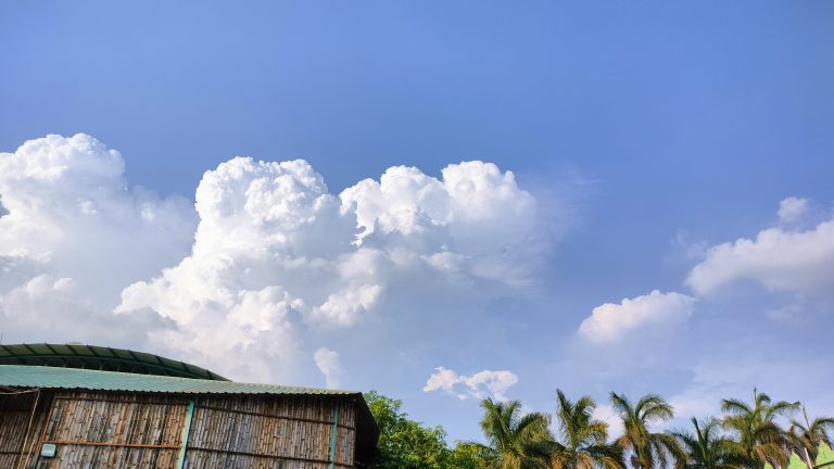 A clear sky adorned with fluffy white clouds over trees and a rooftop.