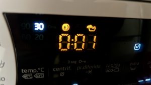 Close-up of a washing machine control panel showing a digital display with "0:01" remaining time. Temperature is set to 30 degrees. Icons for cycle options like spin and delayed start are visible. A blue checkmark is illuminated, indicating an option is selected.