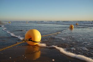 Yellow buoys connected by a rope lie on a sandy beach with gentle waves reaching the shore under a clear blue sky.