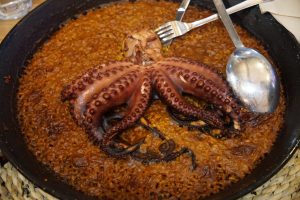 A cooked octopus served on a bed of seasoned rice in a large pan, accompanied by a fork and a spoon.