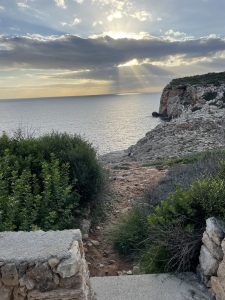 A rocky coastal landscape with a path leading towards the sea at Sant Climent, Menorca. The foreground contains green shrubs and stone steps, while the background features a sun setting over the ocean, casting rays through clouds. 