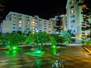 A nighttime view of a residential complex with several multi-story buildings. The buildings have illuminated windows, and there are trees and shrubs in the foreground lit by green lights.