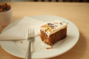 A slice of cake with white frosting, topped with blue and purple edible flowers, seeds, and nuts, sits on a white plate. A fork rests next to it on a wooden table, with a folded napkin beneath the fork.