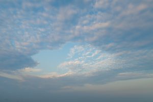 The image shows a peaceful sky, mostly covered with soft clouds. The clouds are light and wispy, with some areas revealing patches of clear blue sky. The lower part of the image transitions into a hazy horizon, with subtle tones of pale blue and gentle hues of pink near the edges of the clouds.