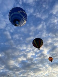 View larger photo: Hot air balloons floating in a cloudy sky, with one large blue and white balloon in the foreground and two smaller, multicolored balloons in the background.