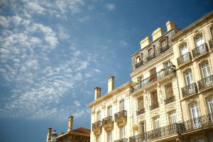 Elegant multi-storey buildings with ornate wrought iron balconies, set against a clear blue sky with scattered clouds. The light stone fa?ade and chimney stacks reflect the bright daylight, giving the architecture a warm and inviting feel.