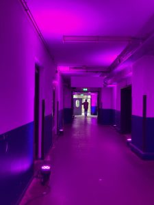 View larger photo: An industrial hallway lit with pink lights. at the end of the hallway there is a door to the bright outside. A person stands in the doorway.