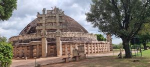 Sanchi Stupa (near Vidisha, (M.P)), India
A large ancient stupa surrounded by intricately carved stone railings and gateways, set in a lush, green landscape. The stupa is made of stone and features detailed carvings, with trees and vegetation visible around the site under a cloudy sky.