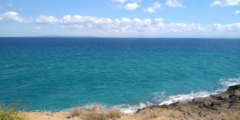 The photo shows majestic cliffs rising above the sea. The water is a deep blue, while the cliffs are covered in natural shades of gray and brown. The sunlight casts gentle shadows, highlighting the texture of the rocks.