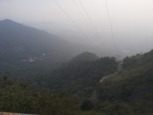 View larger photo: A winding road cuts through a lush, misty mountainous landscape at Western Ghats, Wayanad. Power lines stretch across the scene, and the sky is overcast.