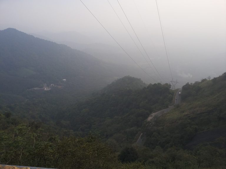 A winding road cuts through a lush, misty mountainous landscape at Western Ghats, Wayanad. Power lines stretch across the scene, and the sky is overcast.