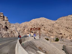 A scenic view of a mountainous landscape with a clear blue sky. The road curves around the rocky terrain, with a sign reading "Jebel Jais Via Ferrata." Two people are walking on the side of the road near a parked white vehicle. Jebel Jais, Ras Al Khaimah