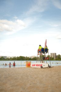 A lifeguard sits on a tall chair overlooking a sandy beach, with a 'Surf Rescue' sign and a rescue board nearby. Swimmers enjoy the water in the background, while beyond the lake, trees and apartment buildings complete the scene under a clear, blue sky.